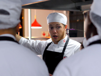 A culinary student in chef whites speaks to classmates in a Swiss Hotel School kitchen